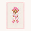 A framed cross-stitch artwork featuring a teddy bear dressed in a white robe with pink accents, including a pink towel on its head and pink slippers. The piece is mounted on a light pink border against an off-white background, with the 'STUDIO daisy' brand logo printed below.