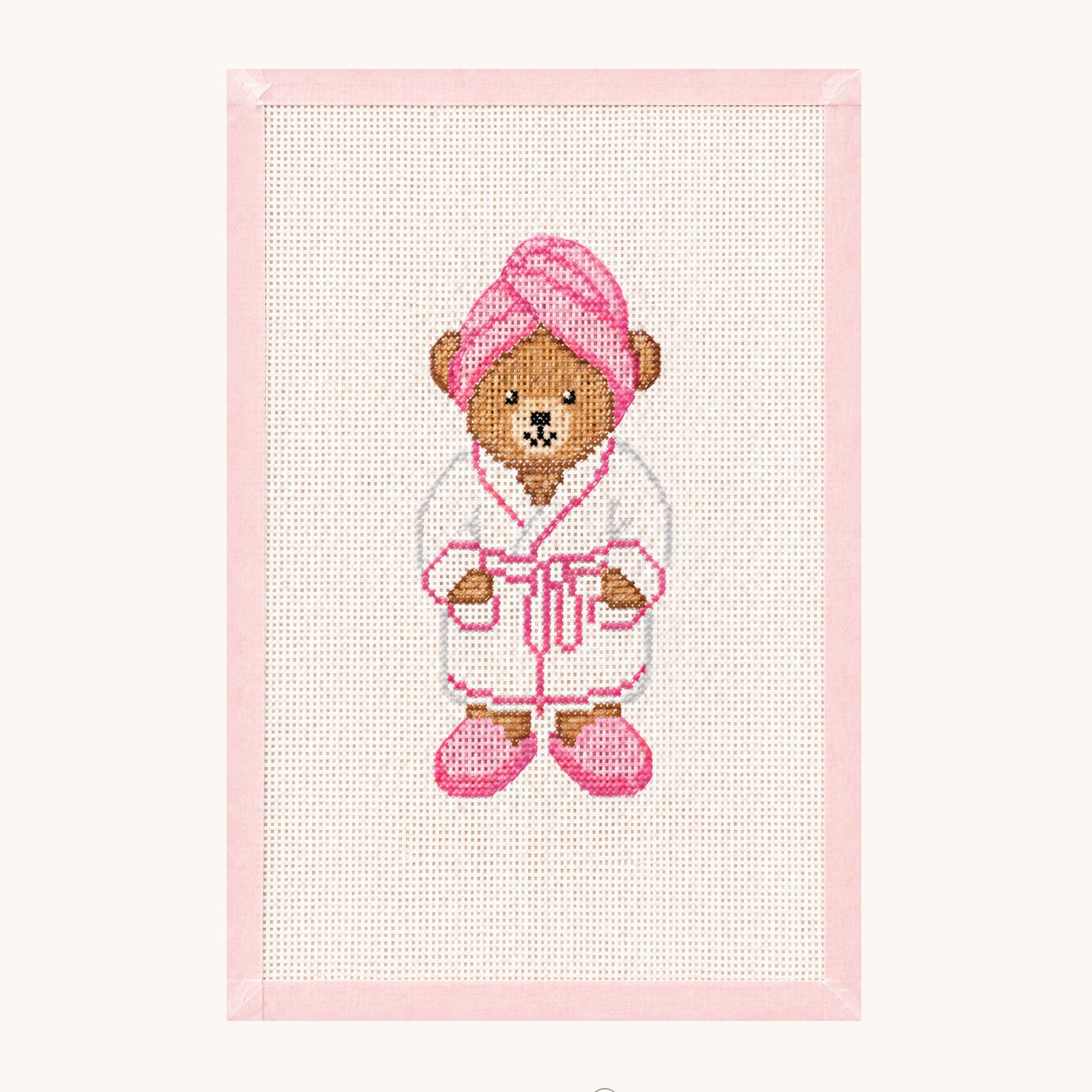A framed cross-stitch artwork featuring a teddy bear dressed in a white robe with pink accents, including a pink towel on its head and pink slippers. The piece is mounted on a light pink border against an off-white background, with the 'STUDIO daisy' brand logo printed below.