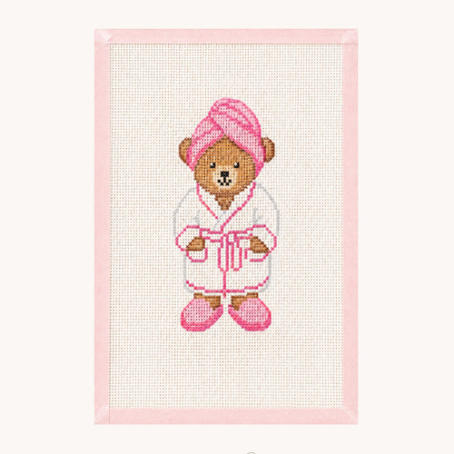 A framed cross-stitch artwork featuring a teddy bear dressed in a white robe with pink accents, including a pink towel on its head and pink slippers. The piece is mounted on a light pink border against an off-white background, with the 'STUDIO daisy' brand logo printed below.
