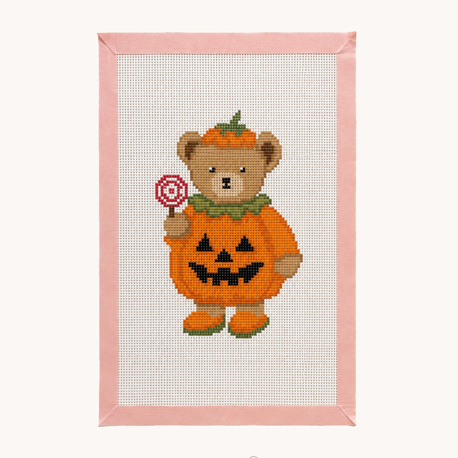 A framed cross-stitch design featuring a teddy bear dressed as a pumpkin holding a lollipop, with a pink stitched border and a white studio background. The brand name 'Studio Daisy' is visible at the bottom.