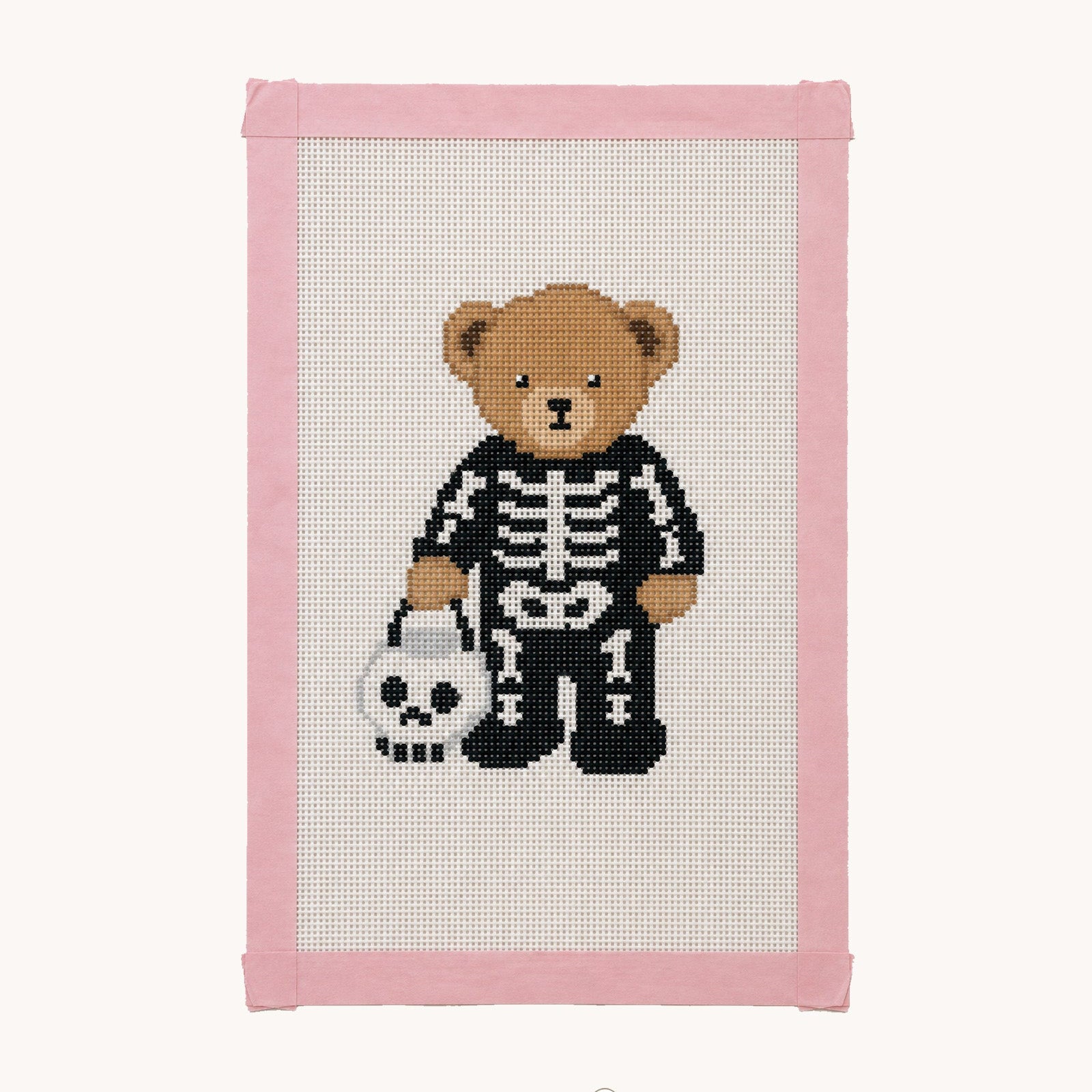 A framed cross-stitch design featuring a teddy bear dressed in a black and white skeleton outfit, holding a white skull bucket. The piece is bordered by a pink fabric frame and displayed against a clean off-white background with a 'Studio Daisy' watermark at the bottom.