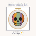 A cross-stitch kit preview showing a colorful skull design stitched inside a wooden embroidery hoop on a light cream background, with the text 'cross-stitch kit' above and 'STUDIO daisy' below.