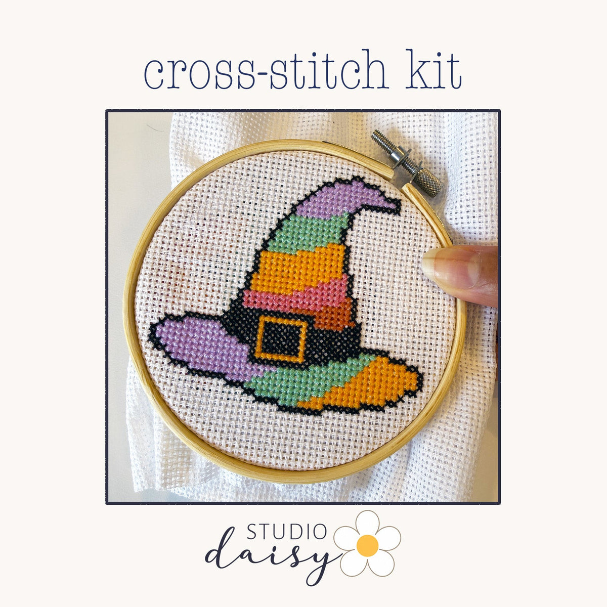 A cross-stitch kit preview showing a completed colorful witch hat design inside a wooden embroidery hoop, with the text 'cross-stitch kit' above and 'STUDIO daisy' below on a light cream background.